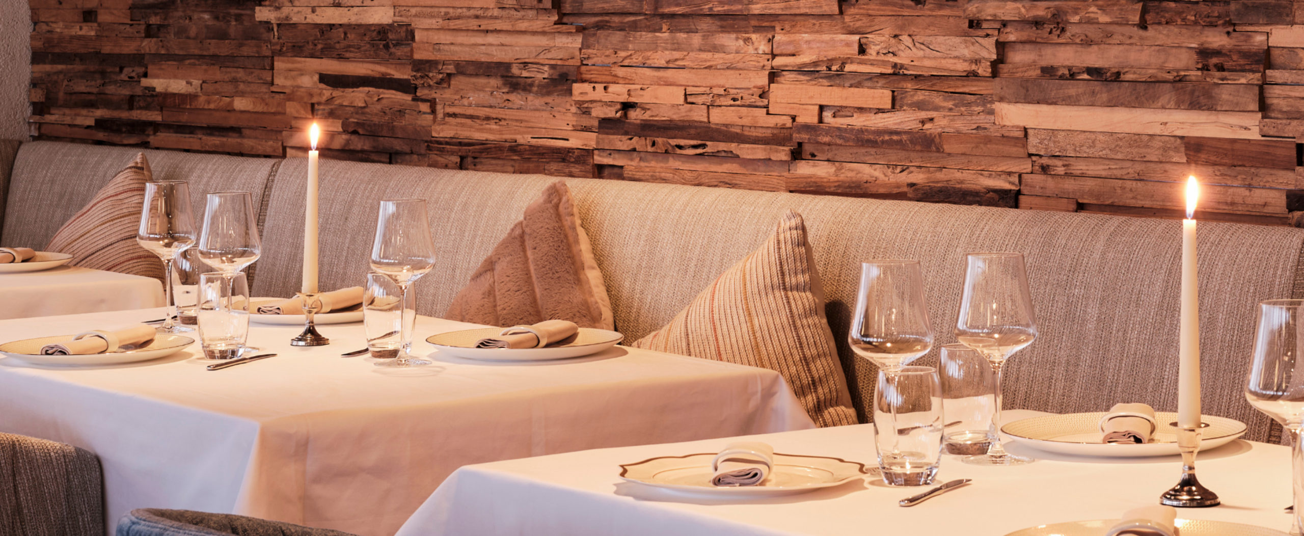 Stilvoll gedeckter Restauranttisch mit Kerzenlicht und Weingläsern vor einer rustikalen Holzwand im Hotel Ambassador Zermatt.