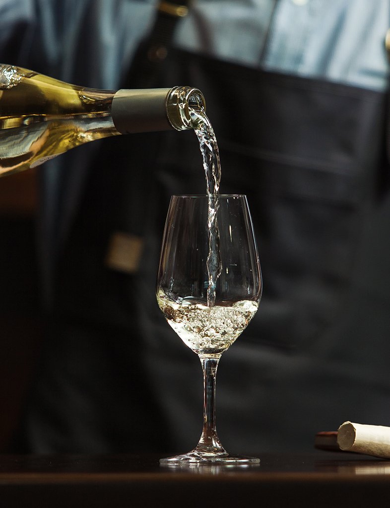 Male sommelier pouring white wine into long-stemmed wineglasses.